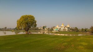 A golf course with a large tree, a body of water, and a building with domed structures in the background. - Gassan Panorama Golf Club (Ban Thi)