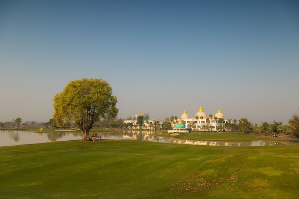 A golf course with a large tree, a body of water, and a building with domed structures in the background.