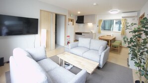 A modern living room with a sofa, a coffee table, a flat-screen TV, and a kitchen area with a dining table and chairs. - Sakura 612 (Sapporo)