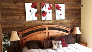 A bedroom with a wooden headboard, a bed with multiple pillows, two bedside tables with lamps, and three wall-mounted paintings of red flowers. - Cody's Happy Trails Ranch Vacation Rental -New Braunfels Texas! (New Braunfels)