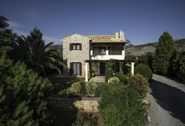 A two-story stone house with a balcony, surrounded by lush greenery and a gravel driveway. - Galaxidi Mansion, Private villa (Galaxidi)