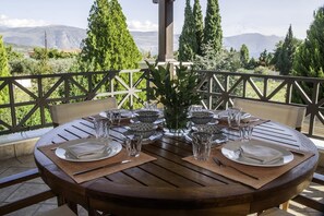 A round wooden table set for four with plates, cutlery, and glasses, surrounded by a white railing with a view of trees and mountains in the background. - Galaxidi Mansion, Private villa (Galaxidi)