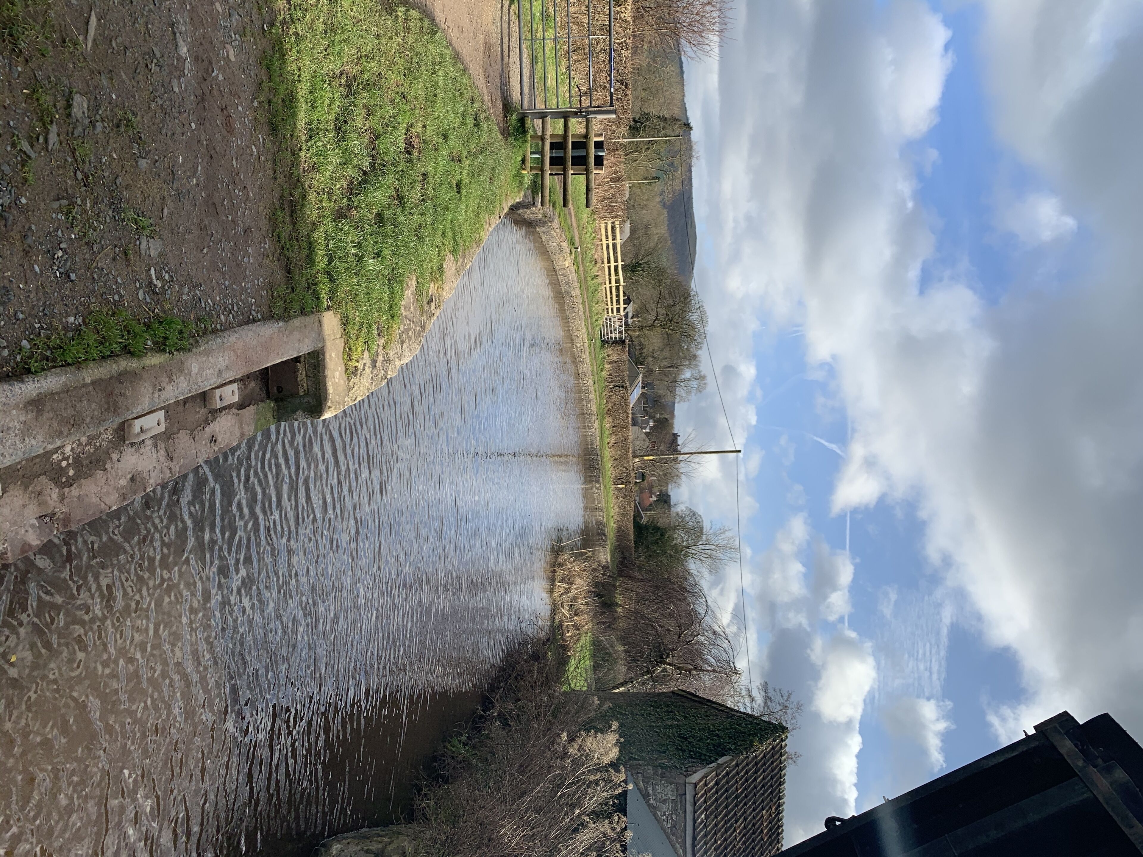 Ein Fluss mit einem Betonweg am Ufer, eine Holzkonstruktion in der Ferne und ein teilweise bewölkter Himmel.