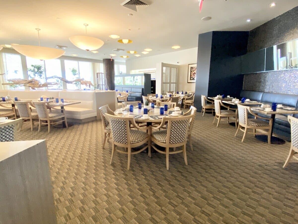A spacious dining area with round tables and chairs, a patterned carpet, and large windows allowing natural light.