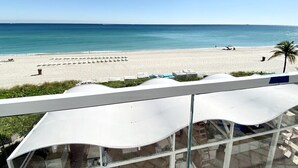 A beach view from a balcony with white canopies, a palm tree, and a clear sky. - Ocean View Suite at T. International STR-02496 (Sunny Isles)