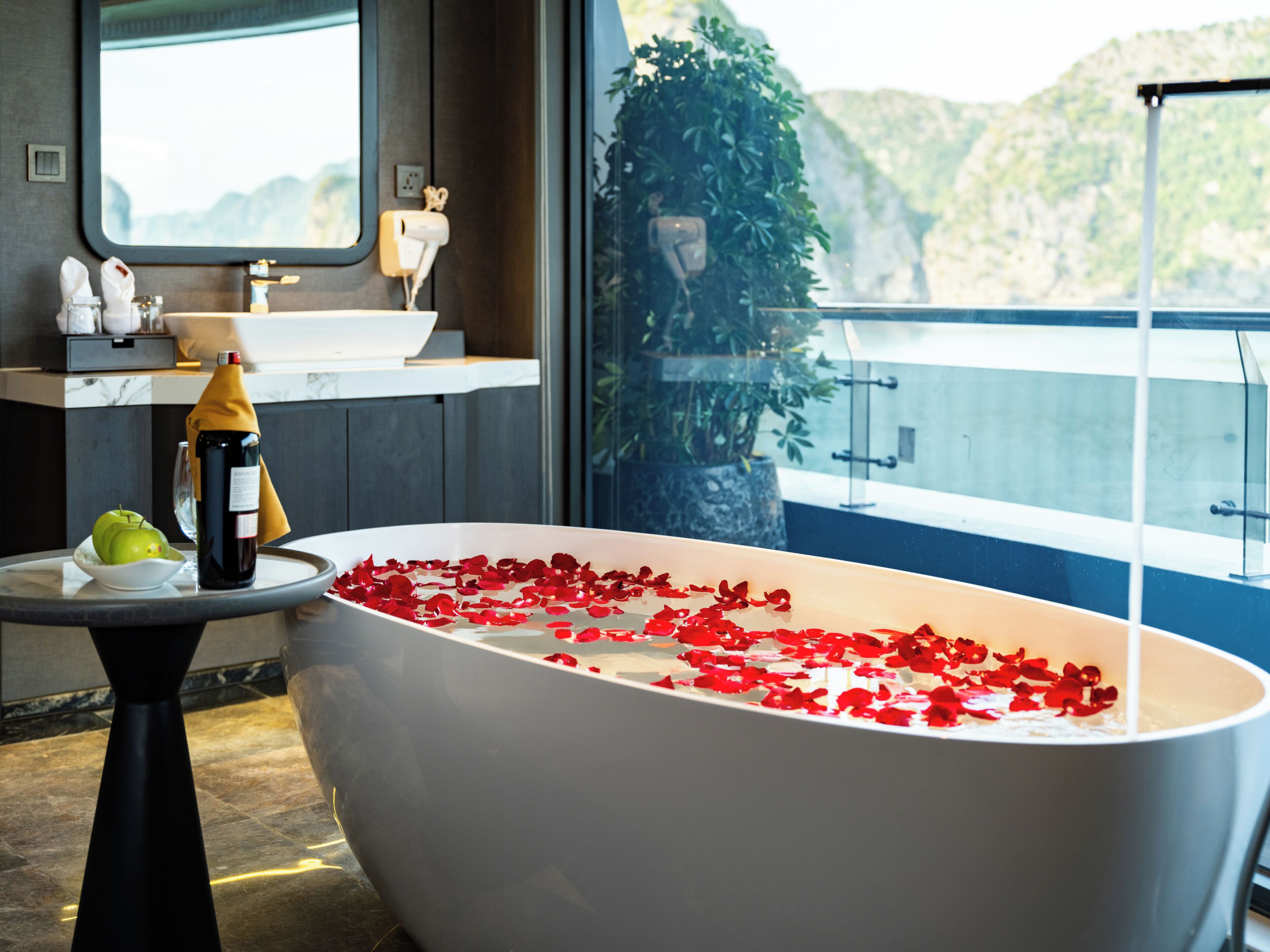 A modern bathroom with a freestanding bathtub filled with rose petals, a bottle of champagne, and a green apple on a marble countertop.