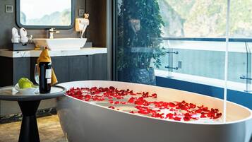 A modern bathroom with a freestanding bathtub filled with rose petals, a bottle of champagne, and a green apple on a marble countertop.