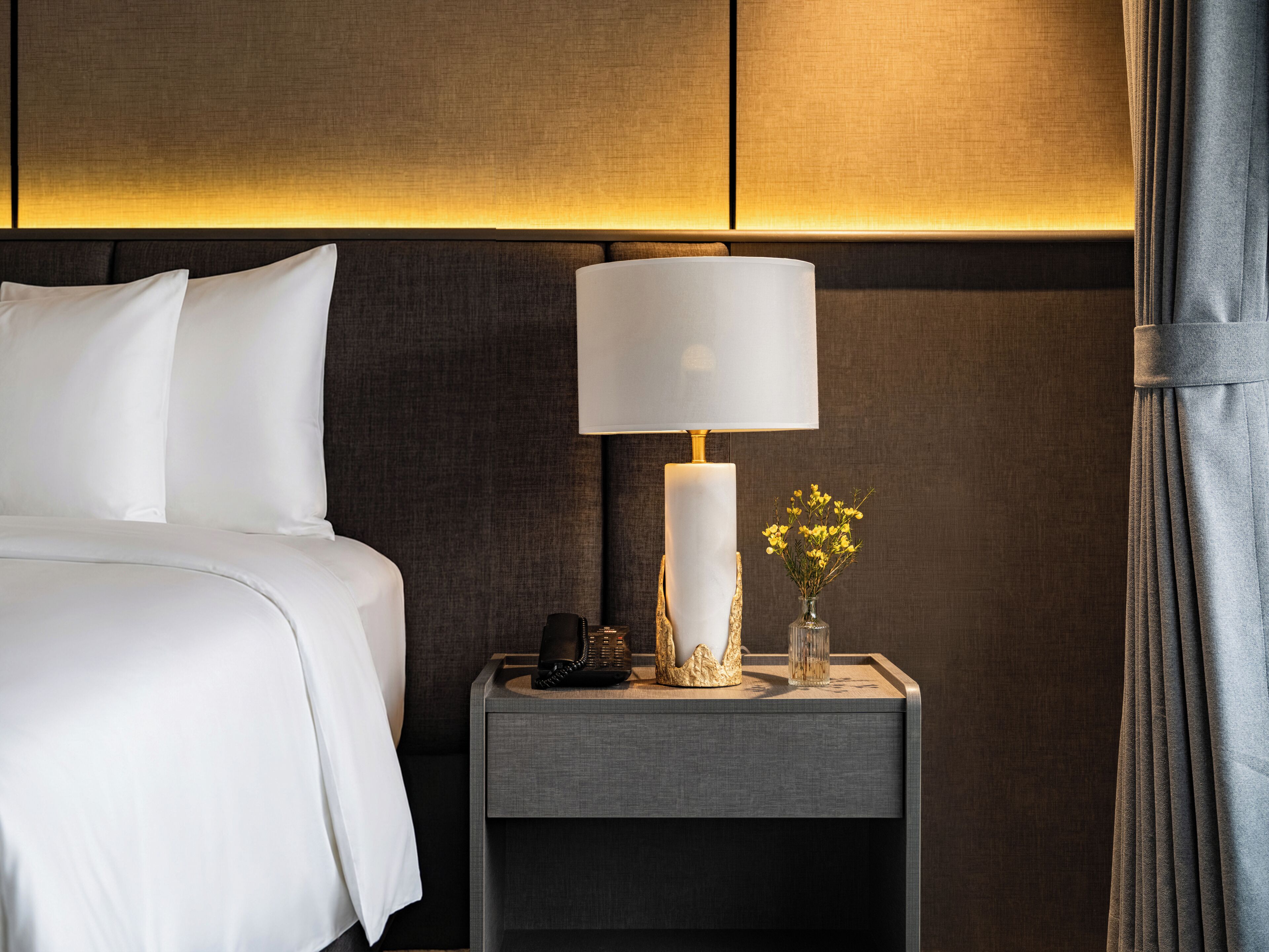Una camera d'albergo con un letto, un comodino con una lampada, un vaso con fiori e un telefono.