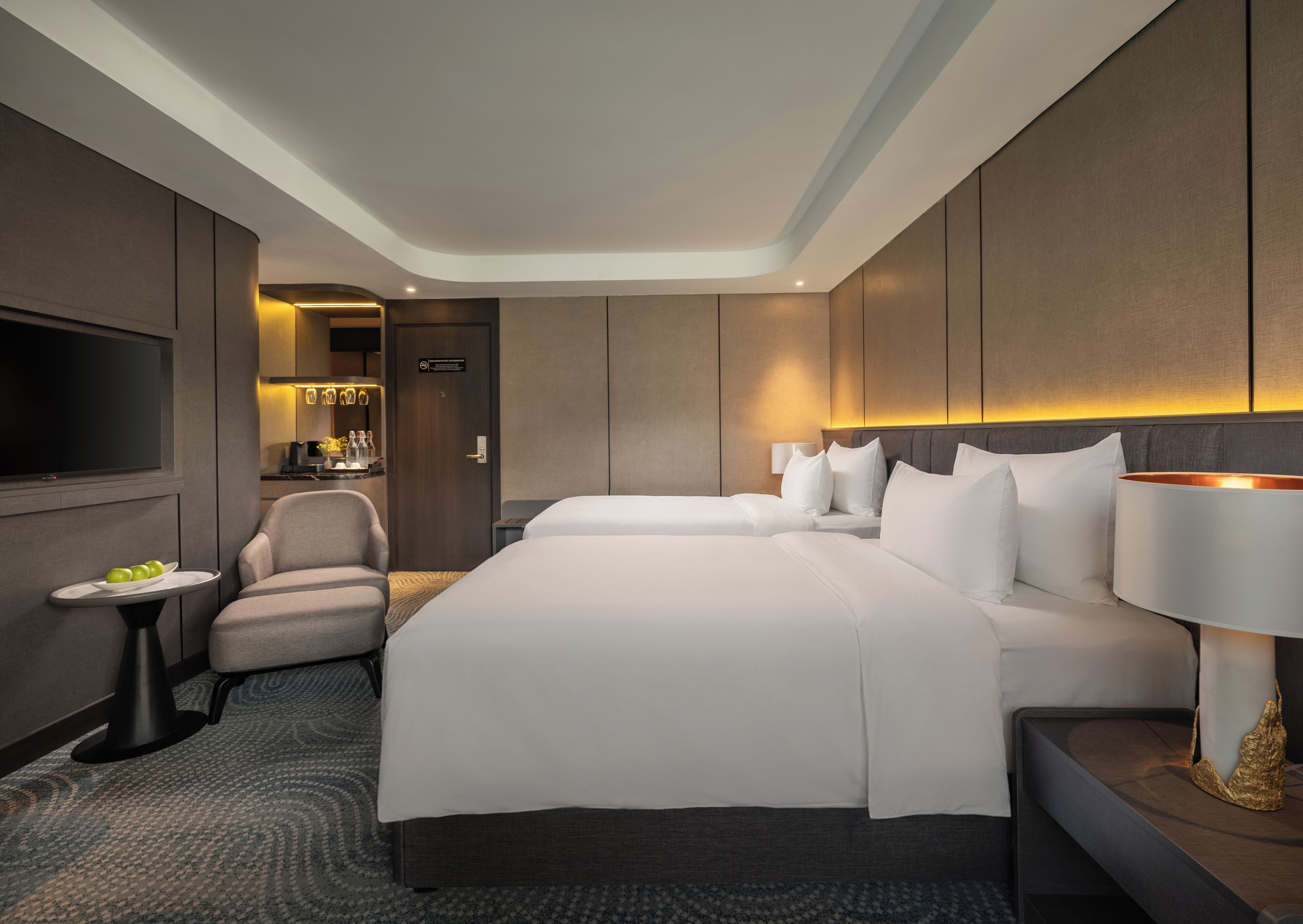 Quarto de hotel com uma cama grande, uma cadeira, uma televisão e uma mesinha com uma tábua de frutas.