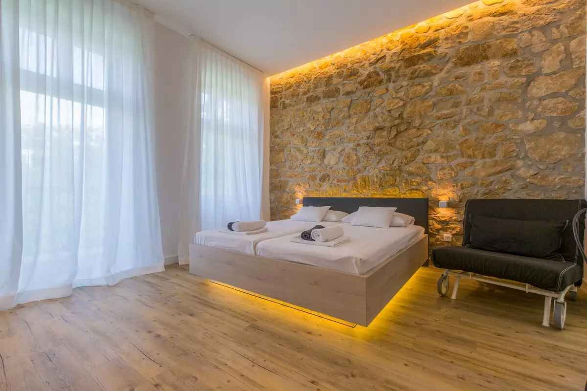 Un dormitorio moderno con una pared de acento de piedra, una cama con ropa de cama blanca, una silla negra y un suelo de madera.