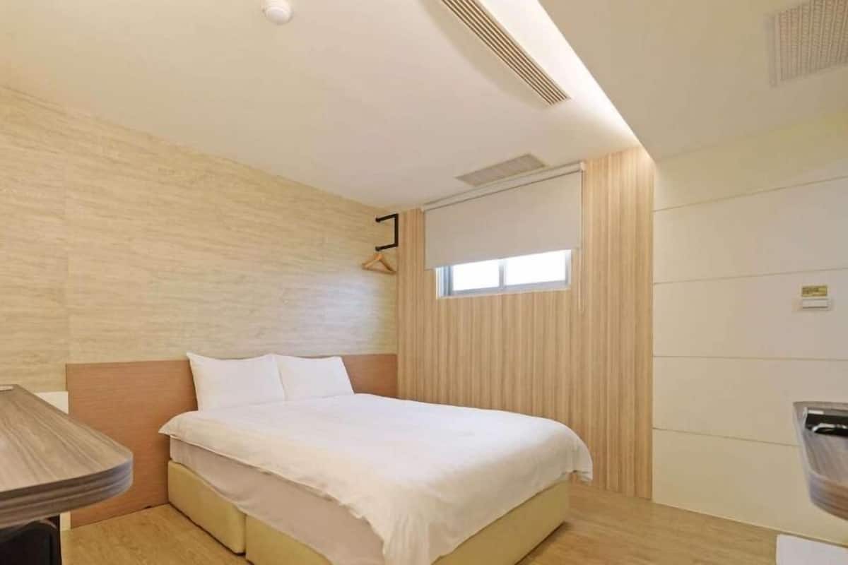 A hotel room with a bed, a bedside table, a hanging light, and a window.