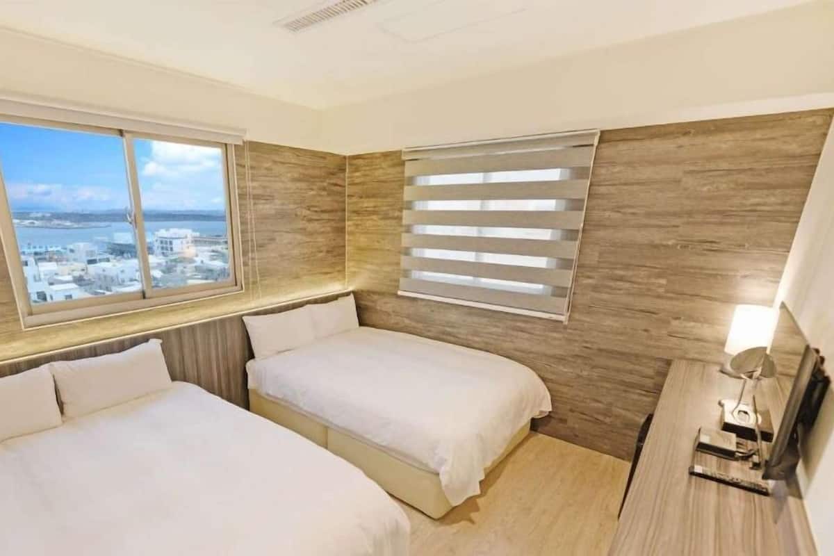 A hotel room with two beds, a large window with a city view, wooden paneling, and a small table with a lamp.