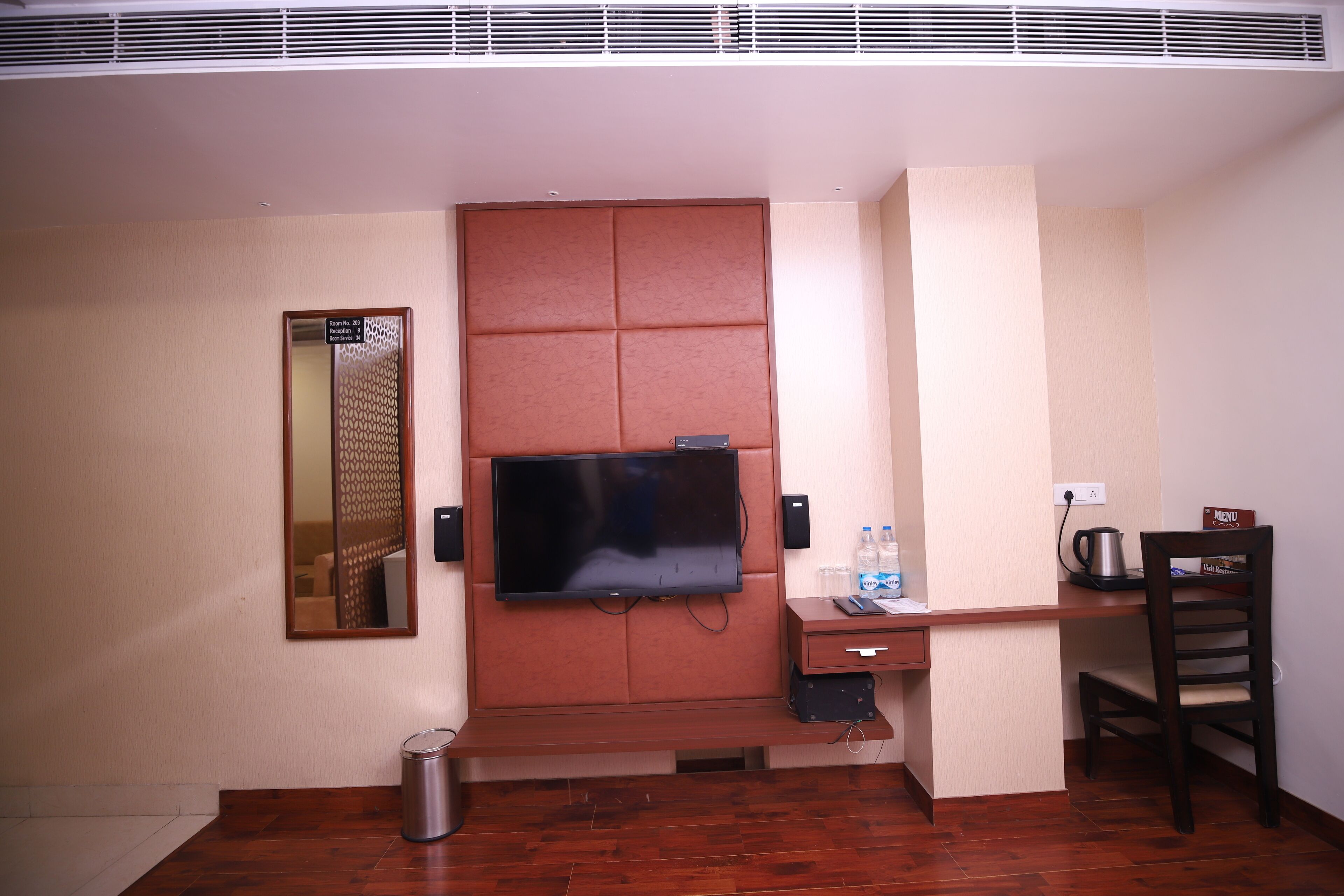 A hotel room with a large flat-screen TV mounted on a wall, a wooden desk with a chair, a trash can, and a small cabinet.