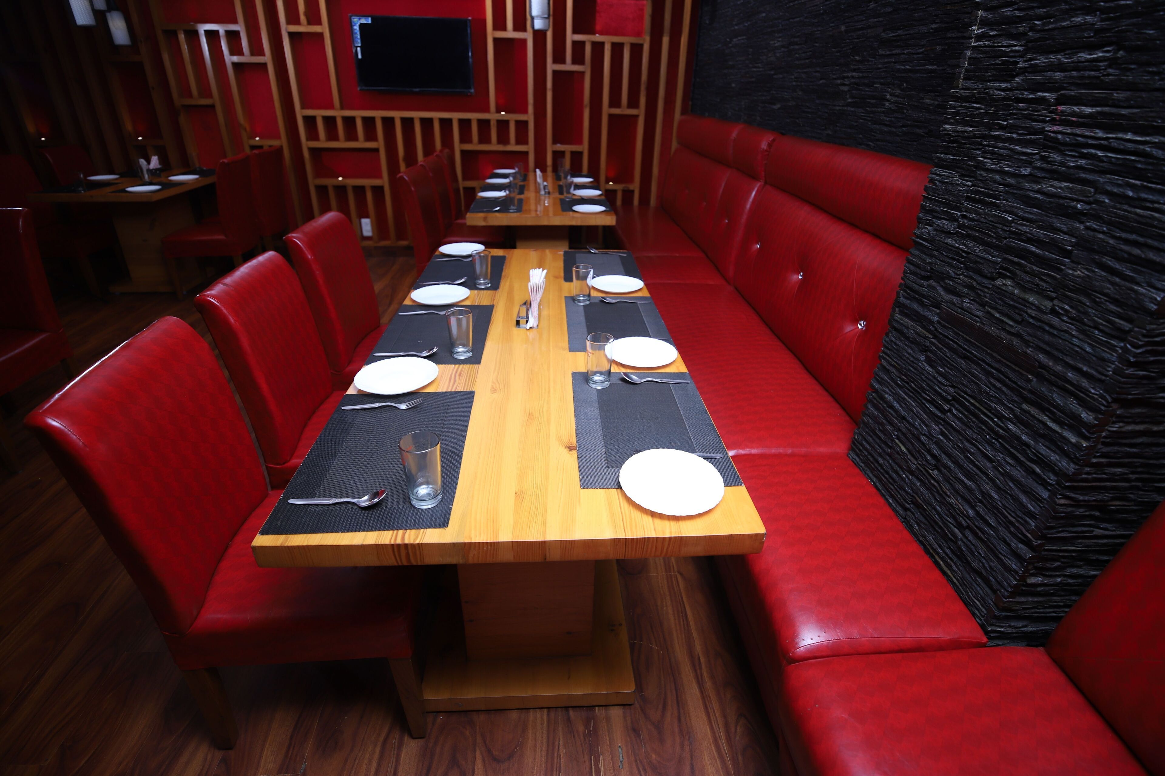 Een restaurant met een lange houten tafel gedekt voor diner, rode bekleden zitjes en een zwart gestructureerde muur.