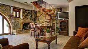 A cozy living room with a stone fireplace, wooden furniture, a spiral staircase, and a television. - Four bedroom Mouri villas complex of 18th century (CPM2+9RH Mouri, Greece)