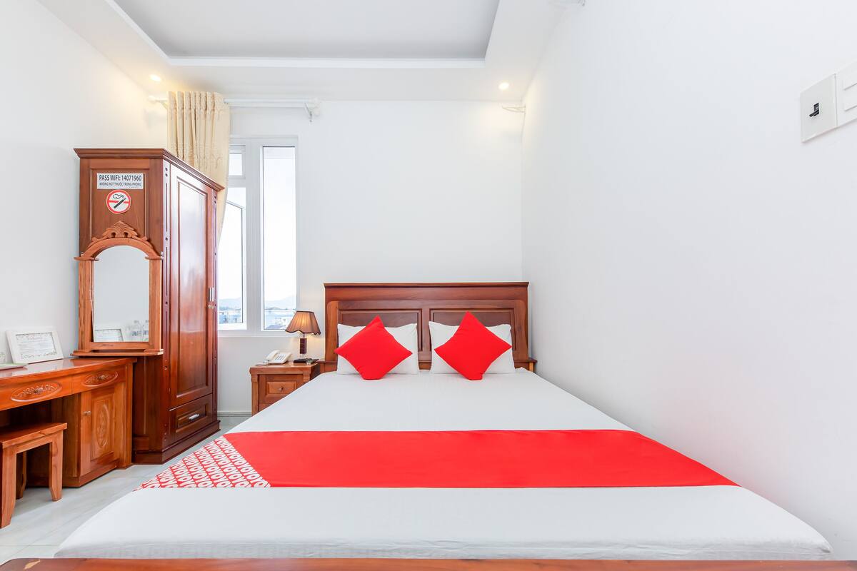 A hotel room with a wooden bed, two red pillows, a nightstand with a lamp, a wooden dresser, and a mirror.
