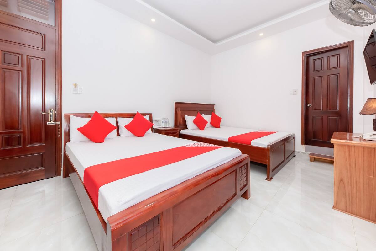 A hotel room with two wooden beds, white bedding, red pillows, a wooden desk, a chair, and a standing fan.