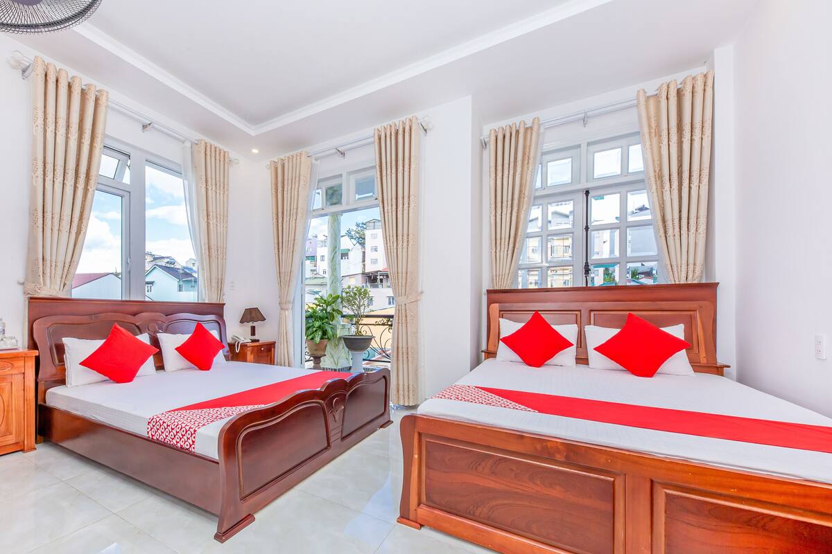 A hotel room with two wooden beds, beige curtains, and a view of buildings through large windows.
