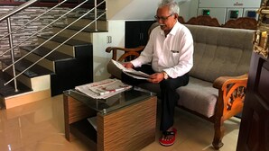 A man sitting on a sofa reading a newspaper. There is a glass coffee table in front of him with a newspaper on it. A staircase with metal railings is visible in the background.