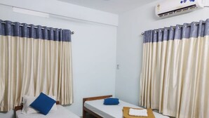 A hotel room with two single beds, each with a wooden frame and a white mattress. There are blue pillows on the beds, and a brown bench in front of one bed. The room has cream-colored curtains, and an air conditioning unit is mounted on the wall above the beds.