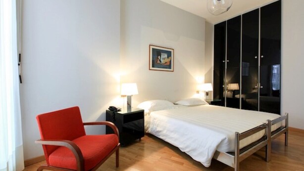 Habitació d'hotel amb un llit, una cadira vermella, una tauleta de nit amb una làmpada, un quadre a la paret i un armari amb portes corredisses.
