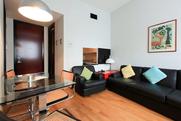 A modern living room with a glass dining table, black leather sofa, and a wooden floor.