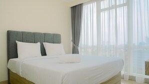 A neatly made bed with white linens and a grey headboard in a bright room with a large window and sheer curtains. - Wonderful 2BR at Menteng Park Apartment (Jakarta)