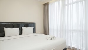 Een net opgemaakt bed met witte lakens en twee opgerolde handdoeken, een donker hoofdbord en dunne gordijnen voor het raam.