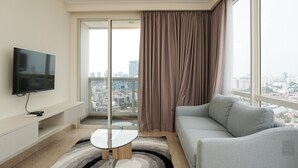 Een moderne woonkamer met een flatscreen televisie, een salontafel met glazen blad, een grijze bank en een uitzicht op een stadslandschap door een groot raam met gordijnen.