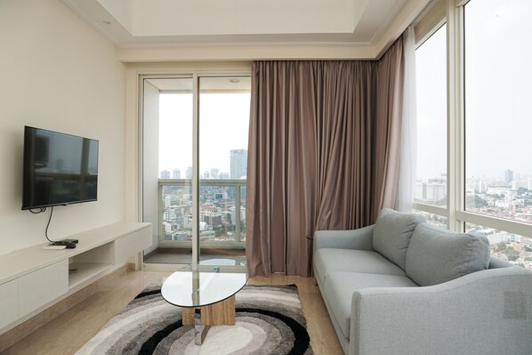 A modern living room with a flat-screen TV, a glass-top coffee table, a grey sofa, and a view of a cityscape through a large window with curtains.
