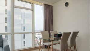 Een moderne eetruimte met een glazen tafel, vier stoelen, een klok aan de muur en uitzicht op gebouwen door grote ramen.