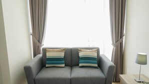A grey sofa with two striped pillows, a lamp on a wooden side table, and beige curtains. - Newly Furnished Studio Apartment at Menteng Park (Jakarta)