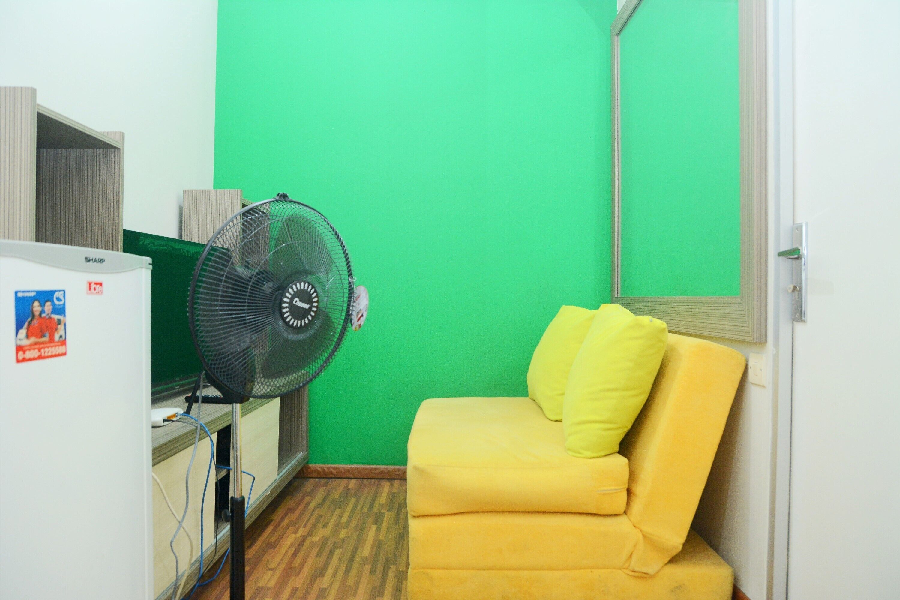 Een kleine kamer met een geel sofa, een groene muur, een witte deur en een zwarte ventilator.
