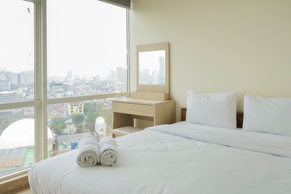 A hotel room with a large window, a bed with white linens, a wooden nightstand, a mirror, and a view of the cityscape.
