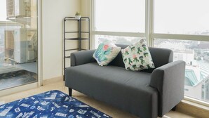 A grey sofa with two patterned pillows, a small black shelf with potted plants, and a view of the city through large windows. - Wonderful 2BR Menteng Park Apartment with Private Lift (Jakarta)
