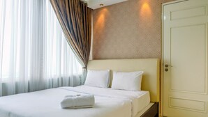 A hotel room with a large bed, white linens, a neatly made white towel, a door, and a window with sheer curtains.