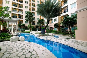 A swimming pool surrounded by a stone pathway, palm trees, and a multi-story building with balconies. - 2BR City Home Apartment (Jakarta)