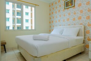 A neatly made bed with white linens and a rolled towel, a window with blinds, a small stool, and a floral wallpaper pattern on the wall. - 2BR City Home Apartment (Jakarta)