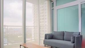 A modern living room with a grey sofa, a wooden coffee table, and large floor-to-ceiling windows offering a view of the cityscape. - Comfortable and Homey 1BR Apartment at Ancol Mansion (Jakarta)
