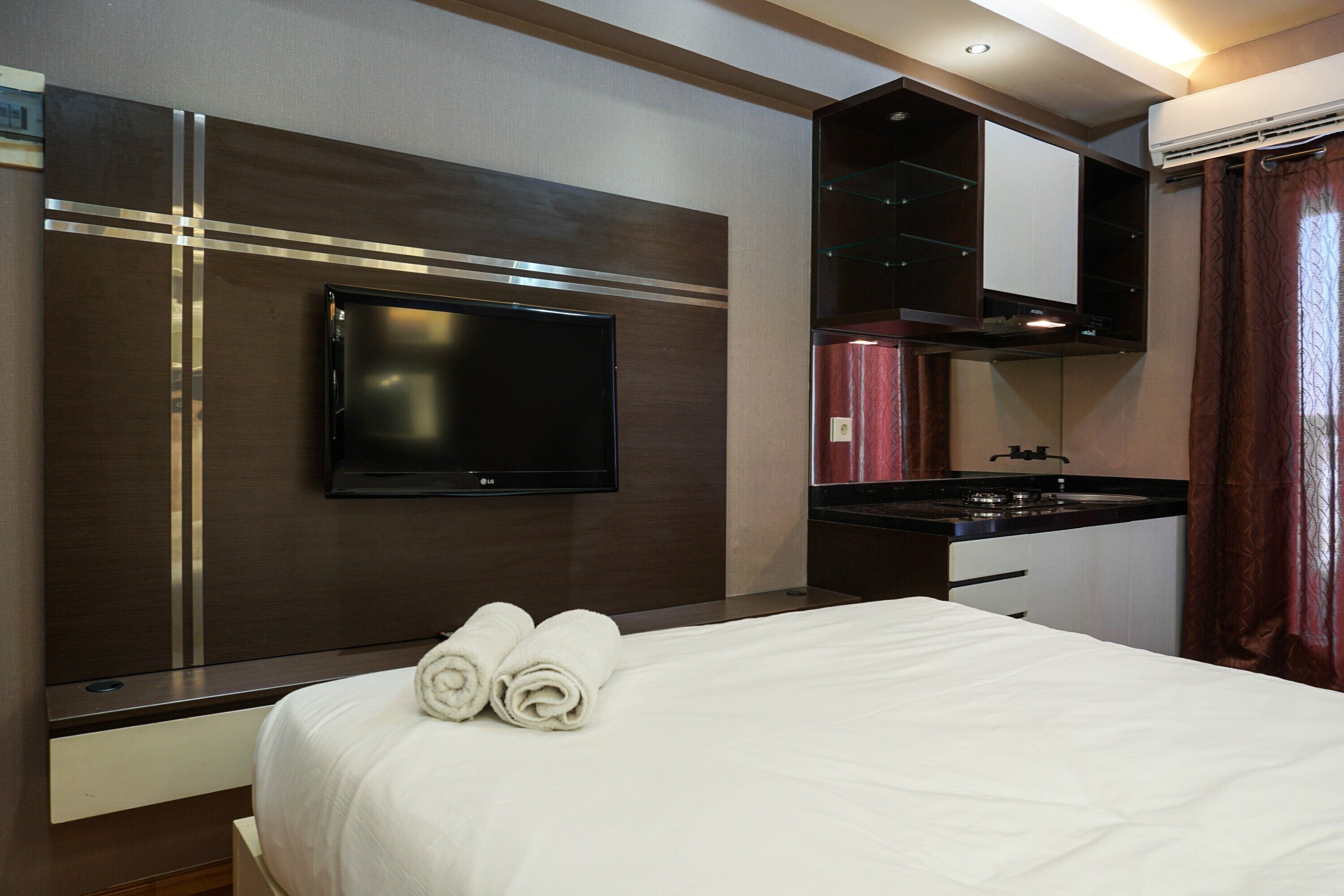 A hotel room with a bed, a flat-screen TV mounted on the wall, a small kitchenette, and a window with curtains.