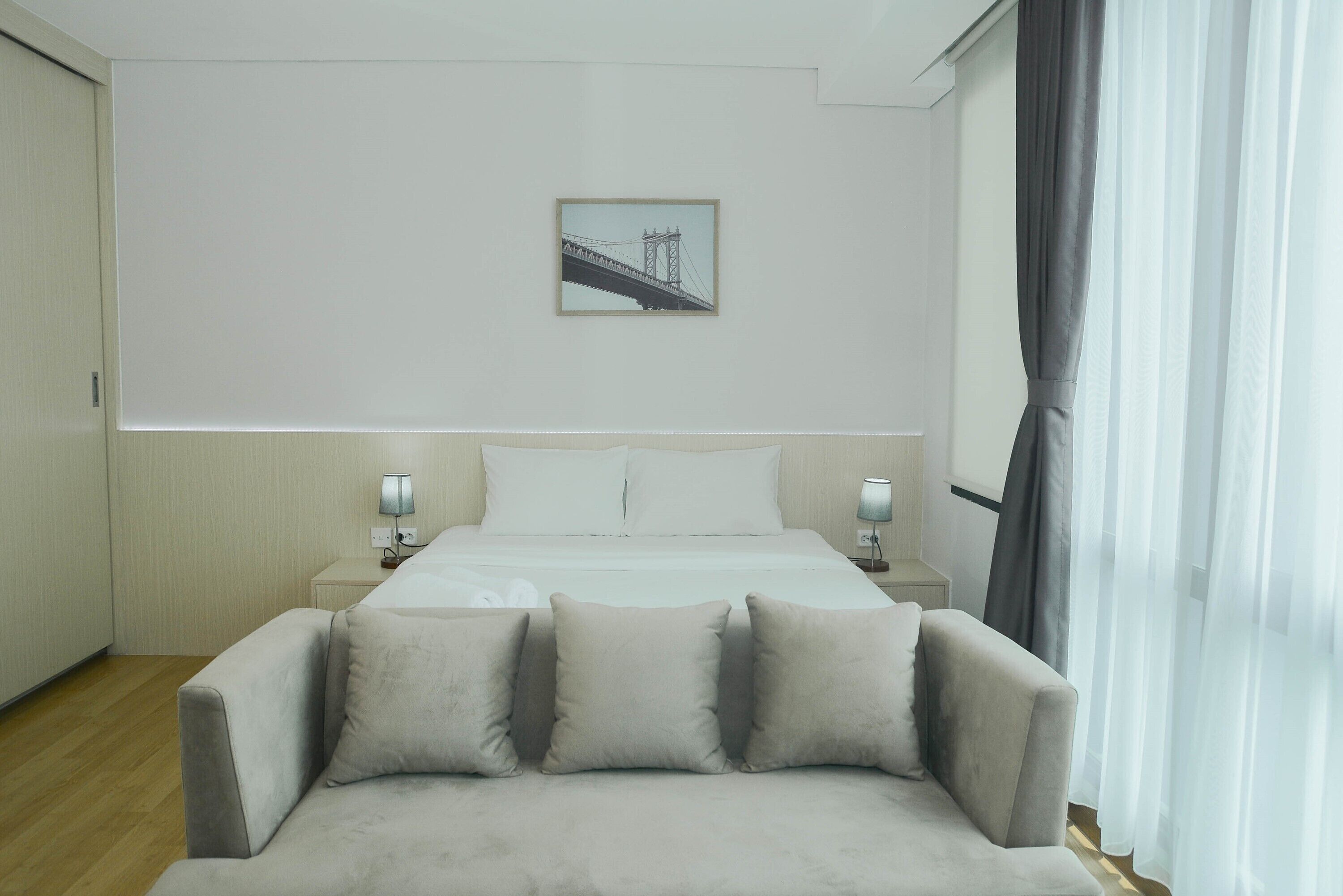 Habitación de hotel con cama, dos mesitas de noche con lámparas, un sofá con cojines, un cuadro en la pared y una ventana con cortinas.