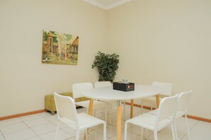 Une pièce avec une table ronde, des chaises blanches, un tableau au mur, une plante verte et un petit banc jaune.
