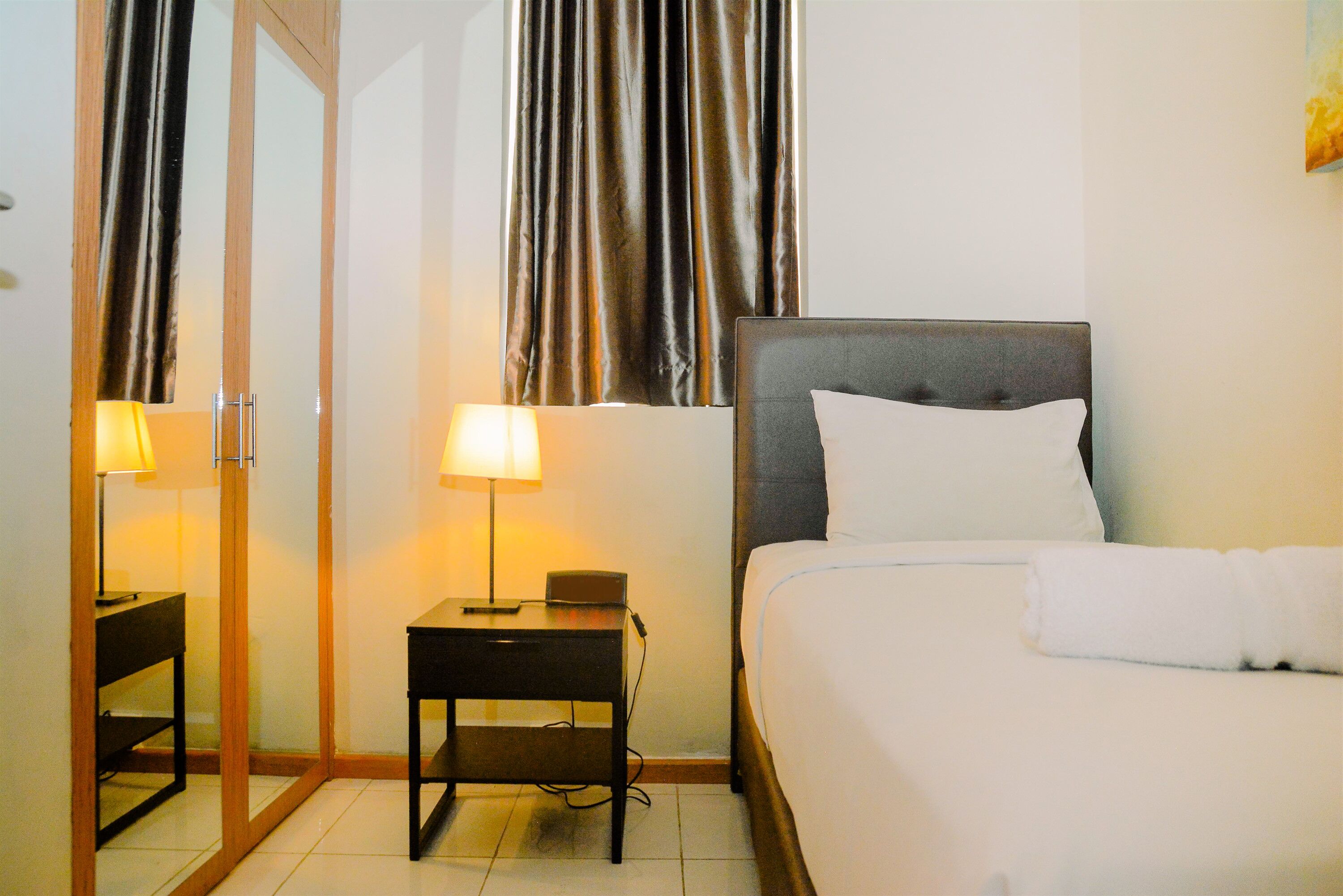 A hotel room with a bed, a nightstand with a lamp, a mirror, and a window with curtains.