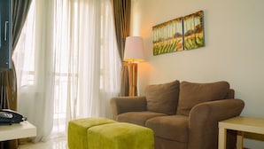 A living room with a brown sofa, a yellow ottoman, a lamp, and a painting on the wall. - 3BR Business Residence at Grand Palace Kemayoran (Jakarta)