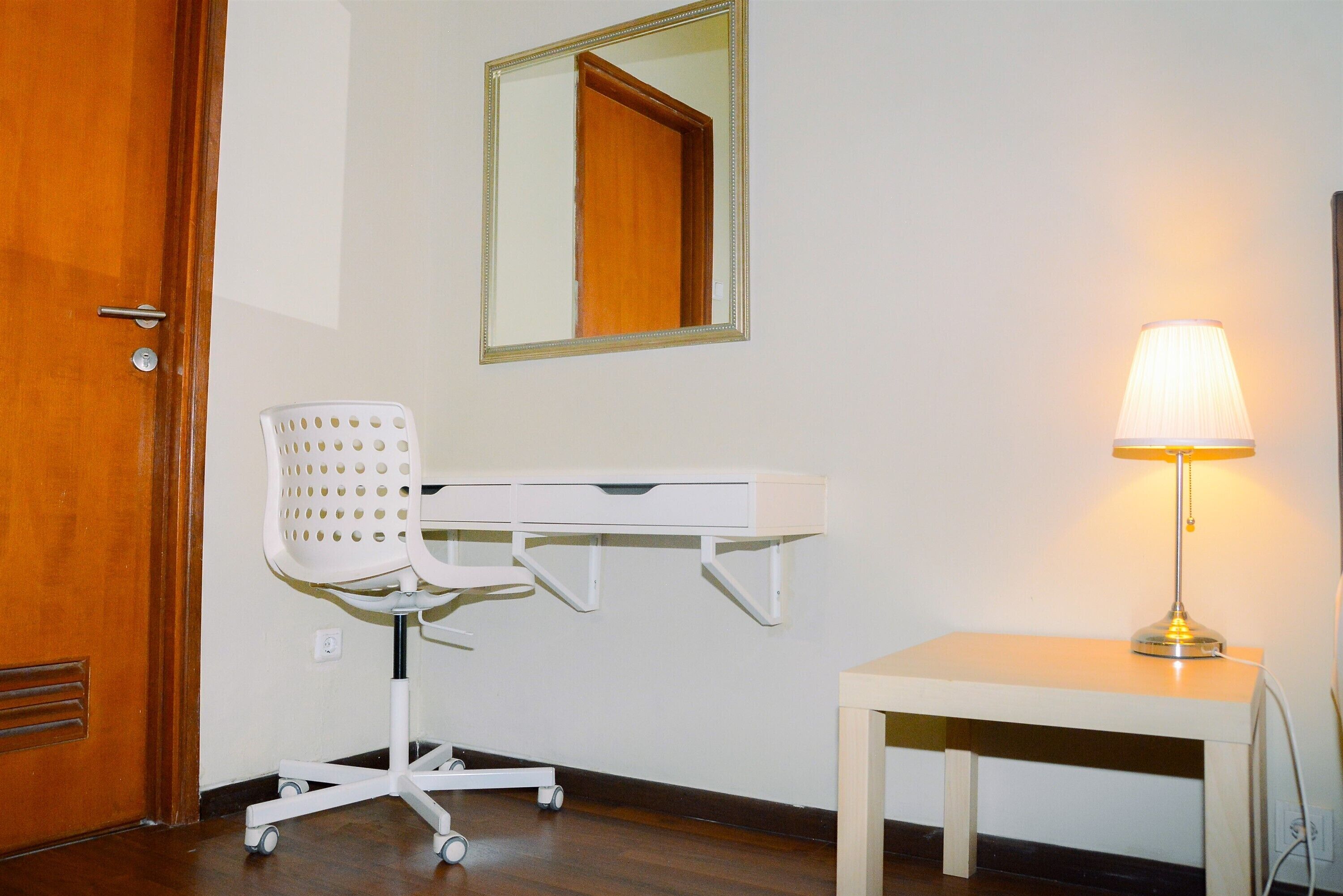 A room with a wooden door, a white desk with drawers, a white office chair, a standing lamp, and a mirror on the wall.