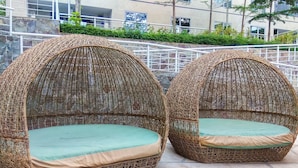 Two wicker-designed outdoor seating areas with cushions, set on a paved surface with a stone wall and greenery in the background. - Spacious 1BR Unit The Mansion Kemayoran Apartment (Jakarta)