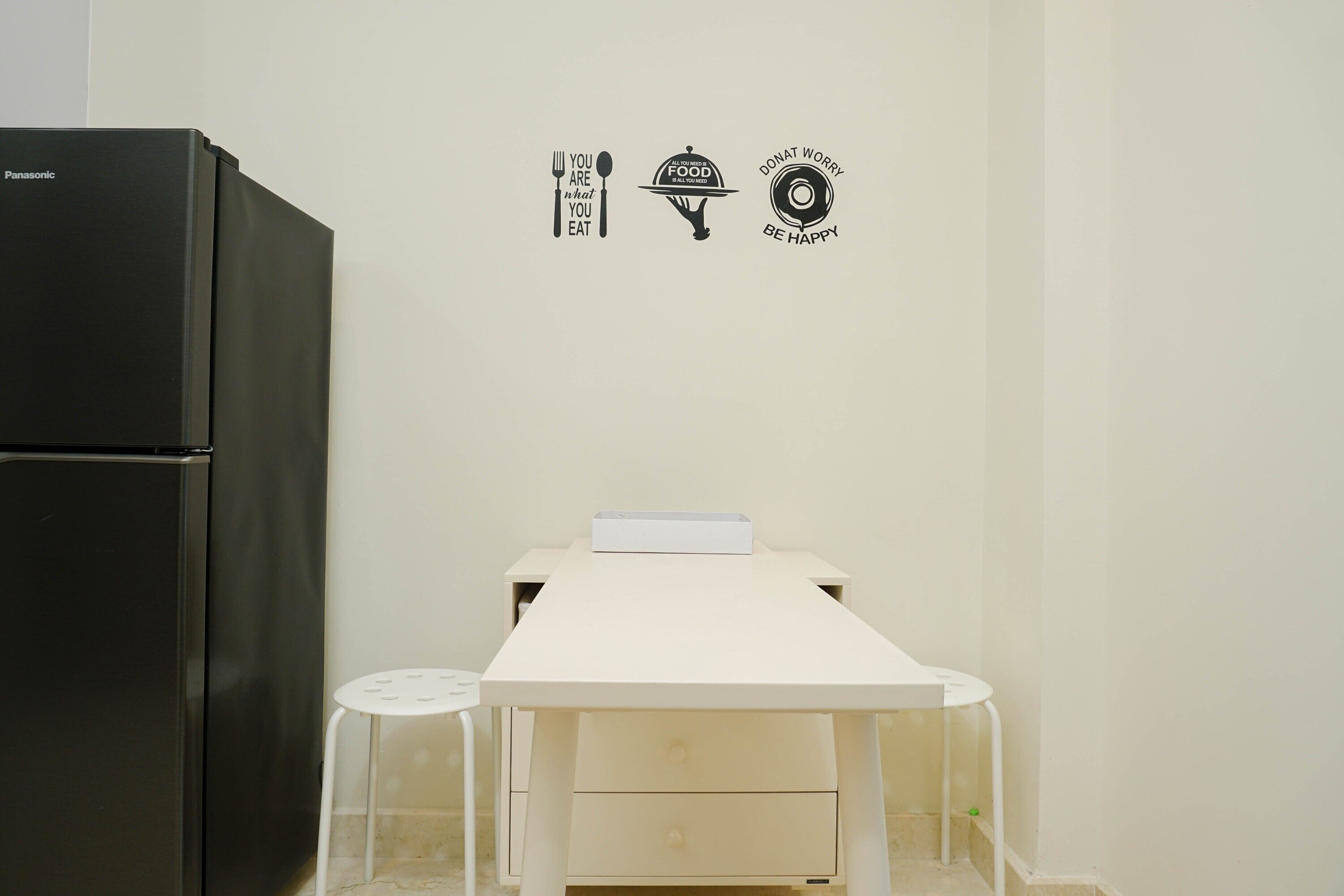 Een keuken met een zwarte koelkast, een witte tafel en een klein wit krukje. Er zijn wandstickers met tekst en afbeeldingen.