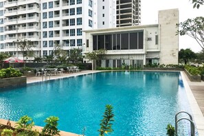 A swimming pool with a wooden deck, surrounded by greenery and a modern building with large windows. - Well Appointed 1BR Apartment at The Aspen Residences (Jakarta)