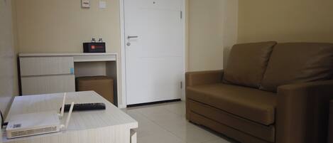 A room with a desk, a brown sofa, a white door, and a small shelf with a travel sign.