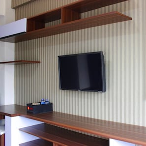 A flat-screen TV mounted on a wall with striped wallpaper, wooden shelves, and a small black box with two water bottles on a wooden table. - Affordable Studio Galeri Ciumbuleuit 2 Apartment near Dago (Bandung)